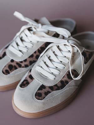 A pair of retro-style lace-up sneakers with cream suede paneling and leopard print fabric insets, shot close-up against a muted mauve background — the kind of shoe that makes a whole outfit without trying.