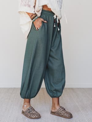 Relaxed harem-style jogger pants in a deep two-tone olive green, styled with a cream lace top, stacked bead bracelets, and leopard print clogs — the kind of outfit that looks effortless but is clearly very intentional.