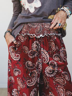 A close-up of wide-leg velvet pants in a deep burgundy paisley print with a smocked waistband, styled with a washed gray appliqué sweatshirt and stacked boho jewelry — the kind of outfit that feels intentional without trying too hard.