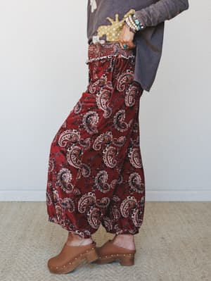 A side-profile shot of wide-leg velvet pants in a rich burgundy paisley print, styled with a washed charcoal top, stacked bracelets, and tan clogs — the kind of outfit that feels intentional without trying too hard.