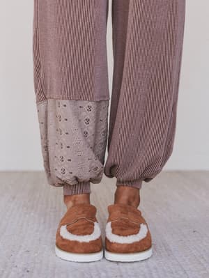 Person wearing brown ribbed sweater and pants with patterned bottom, paired with brown slippers on a light background.