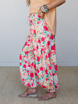 A bold floral harem-style jogger in a vivid red, cream, and green garden print, styled with a peach tee, stacked bracelets, and slide sandals — the kind of outfit that looks effortless but clearly has a point of view.