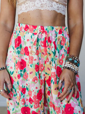 A close-up torso shot of a bold red floral jogger paired with a lace crop top, styled with stacked boho bracelets and silver rings — the kind of outfit that feels like a garden party and a farmers market at the same time.