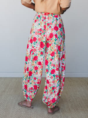 A back-view shot of voluminous floral joggers in a bold red garden print, paired with a peach top and flat sandals — the kind of pants that make a Tuesday feel like a farmer's market morning.