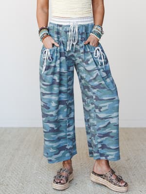 Wide-leg camo pants in a cool-toned blue and green colorway styled with a cream textured crop top, platform espadrille sandals, and stacked beaded bracelets — relaxed and expressive, the kind of outfit that looks effortless without trying.