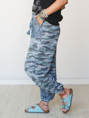 A side-profile shot of blue camo joggers styled with a black graphic tee, turquoise buckle sandals, and a stack of mixed bracelets — relaxed and personal, with lace and tassel details that make the camo feel more boho than utilitarian.