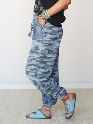 A side-profile shot of blue camo joggers styled with a black graphic tee, turquoise buckle sandals, and a stack of mixed bracelets — relaxed and personal, with lace and tassel details that make the camo feel more boho than utilitarian.