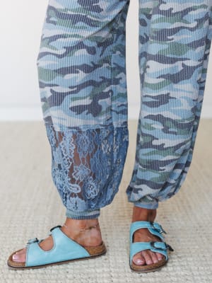 A close-up of blue camo joggers with a flowy lace inset at the hem, styled with light blue buckle sandals on a woven rug — casual and a little unexpected in the best way.
