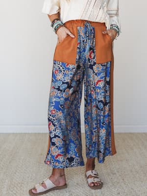 Wide-leg pants in a bold blue floral and paisley print with pumpkin-colored contrast side panels and patch pockets, styled with a cream lace blouse, stacked bracelets, and white buckle sandals — the kind of outfit that looks effortless but clearly wa