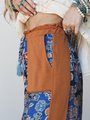 A close-up detail shot of a floral wide-leg pant in cobalt blue and pumpkin, styled with a cream lace top and layered boho jewelry — the contrast patch pocket and tassel tie are the stars here.