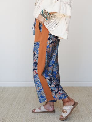 A side-profile lifestyle shot showcasing wide-leg floral pants in deep blue and pumpkin with a contrast side stripe, styled with a cream crochet bell-sleeve top, stacked boho bracelets, and white buckle sandals — the kind of outfit that feels just as