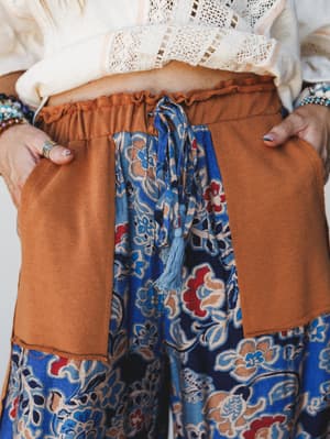 A close-up detail shot of wide-leg pants with a striking color-block design — rust-orange patch pockets and waistband meet a bold blue floral print body, tied together with a tassel drawstring that feels like a small, considered detail worth noticing