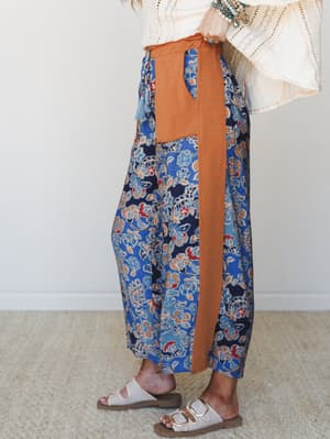 A side-view shot of wide-leg floral pants in navy and cobalt blue with rust orange colorblock paneling and tassel tie detail, styled with a cream lace bell-sleeve top, turquoise jewelry, and white footbed sandals — easy, expressive, and very much the