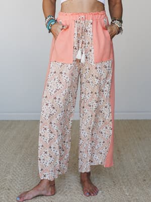 A cropped mid-shot of wide-leg pants in a mixed patchwork design — coral solid panels frame a floral diamond-print center, tied with a tassel drawstring at the waist and styled with layered boho bracelets for a relaxed, lived-in feel.