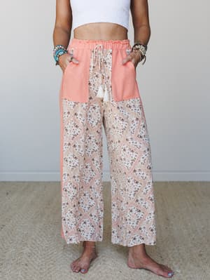 Wide-leg floral patchwork pants in peachy coral and ivory, styled with a white crop top and stacked boho jewelry — the kind of pants that make a grocery run feel like a vacation.