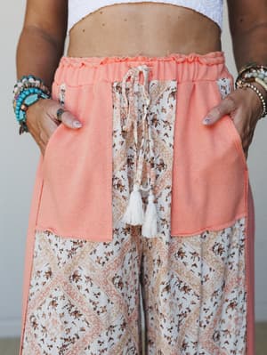 A close-up of wide-leg pants in coral and ivory, styled with hands in pockets — the patchwork floral print and tassel drawstring give them that effortless boho energy that lives somewhere between a beach vacation and a really good farmers market.