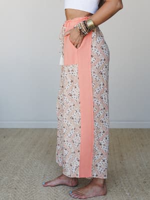 A side-profile shot of wide-leg pants in a floral and medallion print with coral color-block side panels and a frilly elastic waistband, styled with stacked boho bracelets and a white crop top — easy, expressive, and made for barefoot summer days.