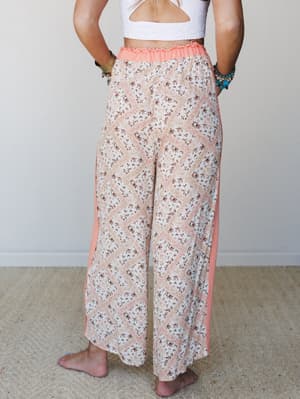 A back-view shot of breezy wide-leg pants in a ditsy floral patchwork print with coral side panels and a smocked waistband, paired with a white textured crop top and stacked bracelets — effortless and a little free-spirited.