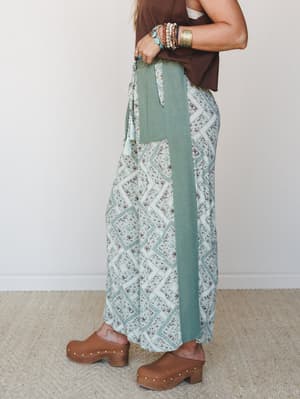 A side-angle lifestyle shot of wide-leg printed pants in sage and cream, styled with a chocolate brown crop tank, wood-studded clogs, and a full stack of turquoise and gold jewelry — the kind of outfit that looks effortless but was absolutely thought