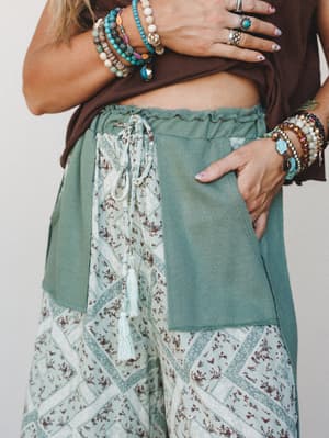 A close-up detail shot of mixed-fabric wide-leg pants in sage and reseda green, featuring a floral patchwork print, tassel drawstring, and ruffle waistband — styled with a brown cropped tee and stacked boho bracelets for a collected, earthy feel.