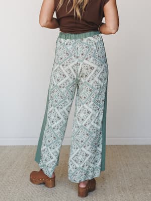 Shot from behind, a wide-leg pant in a patchwork-style floral print takes center stage — paired with a chocolate brown crop tee and wood-soled clogs, the whole look has that effortless, vintage-market energy.