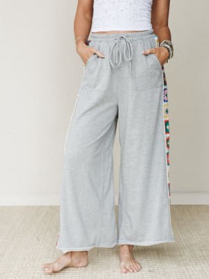 Heather gray wide-leg drawstring pants with colorful granny square crochet side panels, styled with a white textured crop top and stacked bracelets — comfy and expressive in equal measure.