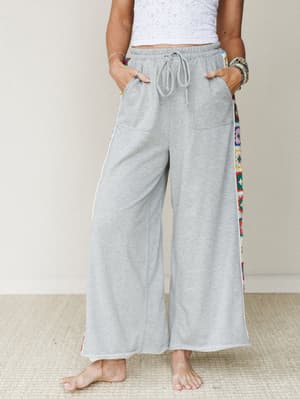 Heather gray wide-leg drawstring pants with colorful granny square crochet side panels, styled with a white textured crop top and stacked bracelets — comfy and expressive in equal measure.