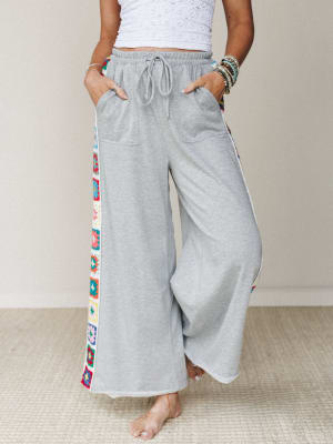 Wide-leg heather gray sweatpants with colorful granny square crochet side panels, styled casually with a white cropped top and stacked bracelets — comfy confidence with a handmade, boho twist.