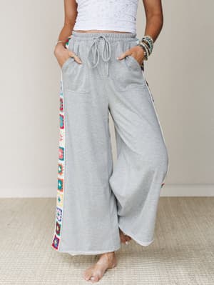 Wide-leg heather gray sweatpants with colorful granny square crochet side panels, styled casually with a white cropped top and stacked bracelets — comfy confidence with a handmade, boho twist.