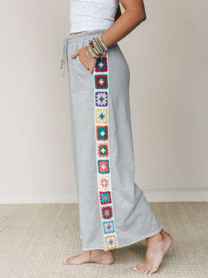 Heather gray wide-leg sweatpants styled with a white lace crop top, the real moment being a vertical strip of colorful granny square crochet running down the outer seam — cozy basics with a handmade, boho soul.