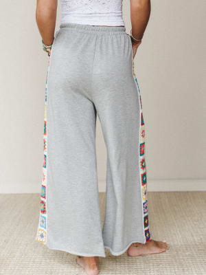 Heather gray wide-leg sweatpants shot from behind, the real story running down both sides — colorful granny square crochet panels that turn a cozy staple into something worth turning around for.