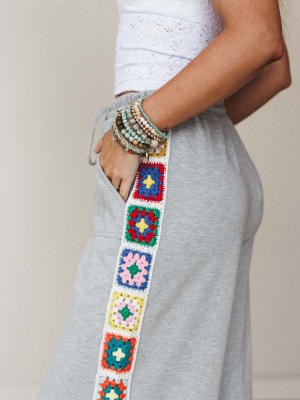 A close-up side shot of heather gray wide-leg sweatpants featuring a vertical granny square crochet panel running down the leg, styled with a white eyelet crop top and a stacked beaded bracelet set — casual and expressive in equal measure.