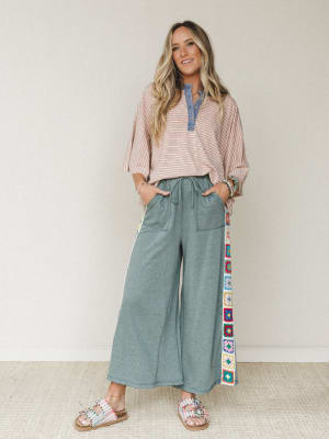 A wide-leg cropped pant in heathered sage with a colorful granny square crochet side panel steals the show, styled with an oversized striped henley top and embellished sandals against a clean studio wall.
