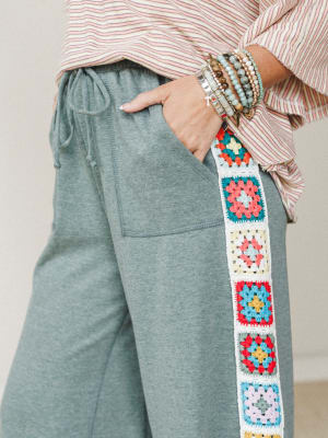 A close-up of heathered sage jogger pants with a vertical granny square crochet panel running down the side seam, styled with a striped top and a stack of beaded bracelets — the kind of outfit that feels intentional without trying too hard.