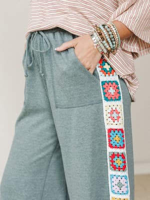 A close-up of heathered sage jogger pants with a vertical granny square crochet panel running down the side seam, styled with a striped top and a stack of beaded bracelets — the kind of outfit that feels intentional without trying too hard.