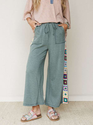 Wide-leg pants in heathered sage green take center stage here, with a vertical strip of colorful granny square crochet running down the side seam — the kind of detail that makes a relaxed pant feel genuinely expressive, styled with a striped oversize