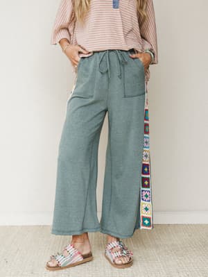 Wide-leg pants in heathered sage green take center stage here, with a vertical strip of colorful granny square crochet running down the side seam — the kind of detail that makes a relaxed pant feel genuinely expressive, styled with a striped oversize