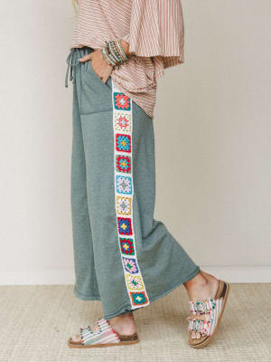 A side-profile shot of wide-leg pants in heathered sage green, featuring a colorful granny square crochet panel running down the outer seam — styled with a red-stripe top, stacked bracelets, and boho sandals for a look that's equal parts cozy and exp