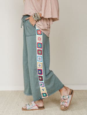 A side-profile shot of wide-leg pants in heathered sage green, featuring a colorful granny square crochet panel running down the outer seam — styled with a red-stripe top, stacked bracelets, and boho sandals for a look that's equal parts cozy and exp