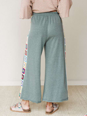 A back-view shot of wide-leg fleece pants in heathered sage, detailed with colorful granny square crochet panels running down each side seam — cozy enough for the couch, interesting enough to actually leave the house.