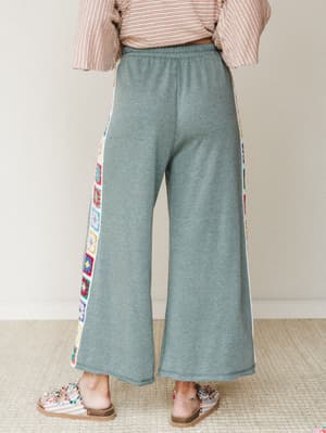 A back-view shot of wide-leg fleece pants in heathered sage, detailed with colorful granny square crochet panels running down each side seam — cozy enough for the couch, interesting enough to actually leave the house.