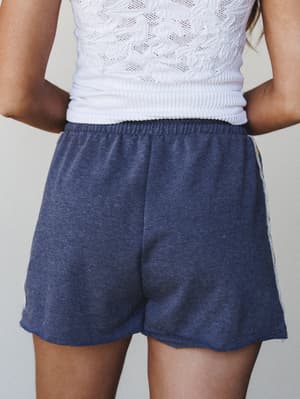 A back-view close-up of heather navy pull-on shorts with a curved raw hem, paired with a white crochet top — casual and comfortable with a relaxed, lived-in feel.