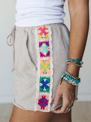 A close-up of relaxed oatmeal shorts with a vertical granny square crochet panel running down the side seam — multicolor and handcraft-y in the best way — paired with a white ribbed top and a full stack of beaded bracelets.