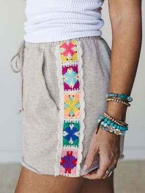 A close-up of relaxed oatmeal shorts with a vertical granny square crochet panel running down the side seam — multicolor and handcraft-y in the best way — paired with a white ribbed top and a full stack of beaded bracelets.