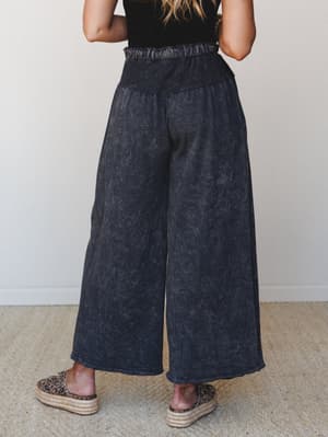 Shot from behind, this model shows off a pair of wide-leg charcoal pants with a lived-in mineral wash texture and smocked high waist — paired with a black crochet top and leopard print platform espadrilles for an effortlessly expressive look.