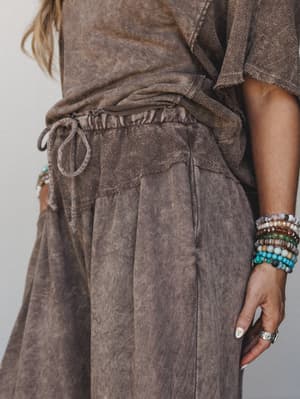 A close-up detail shot of wide-leg mocha pants with a washed, lived-in texture, styled with a knotted tee and a stacked arm of colorful beaded bracelets — the kind of effortless combo that looks like it took zero effort and exactly the right amount.