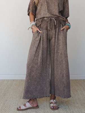 A mocha acid-wash wide-leg pant styled as a matching set with a relaxed tee, hands tucked in pockets, layered bead bracelets and slide sandals giving it that effortless, lived-in weekend feel.