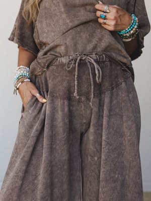 A close-up mid-shot of a mineral-washed wide-leg pant in mocha brown, styled with a matching top and layered boho jewelry — stacked beaded bracelets and silver statement rings bring the whole earthy, effortless look together.