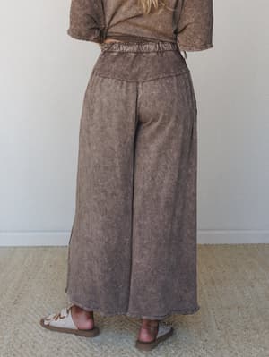 A back-view shot of wide-leg pants in a rich mocha mineral wash, paired with a matching cropped tee and sandals — the kind of effortless set that looks pulled-together without trying.