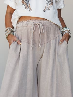 A close-up mid-shot of wide-leg taupe pants with a layered terry-textured waistband and drawstring tie, styled with a cropped graphic tee and stacked boho bracelets — the kind of outfit that feels effortless without trying.